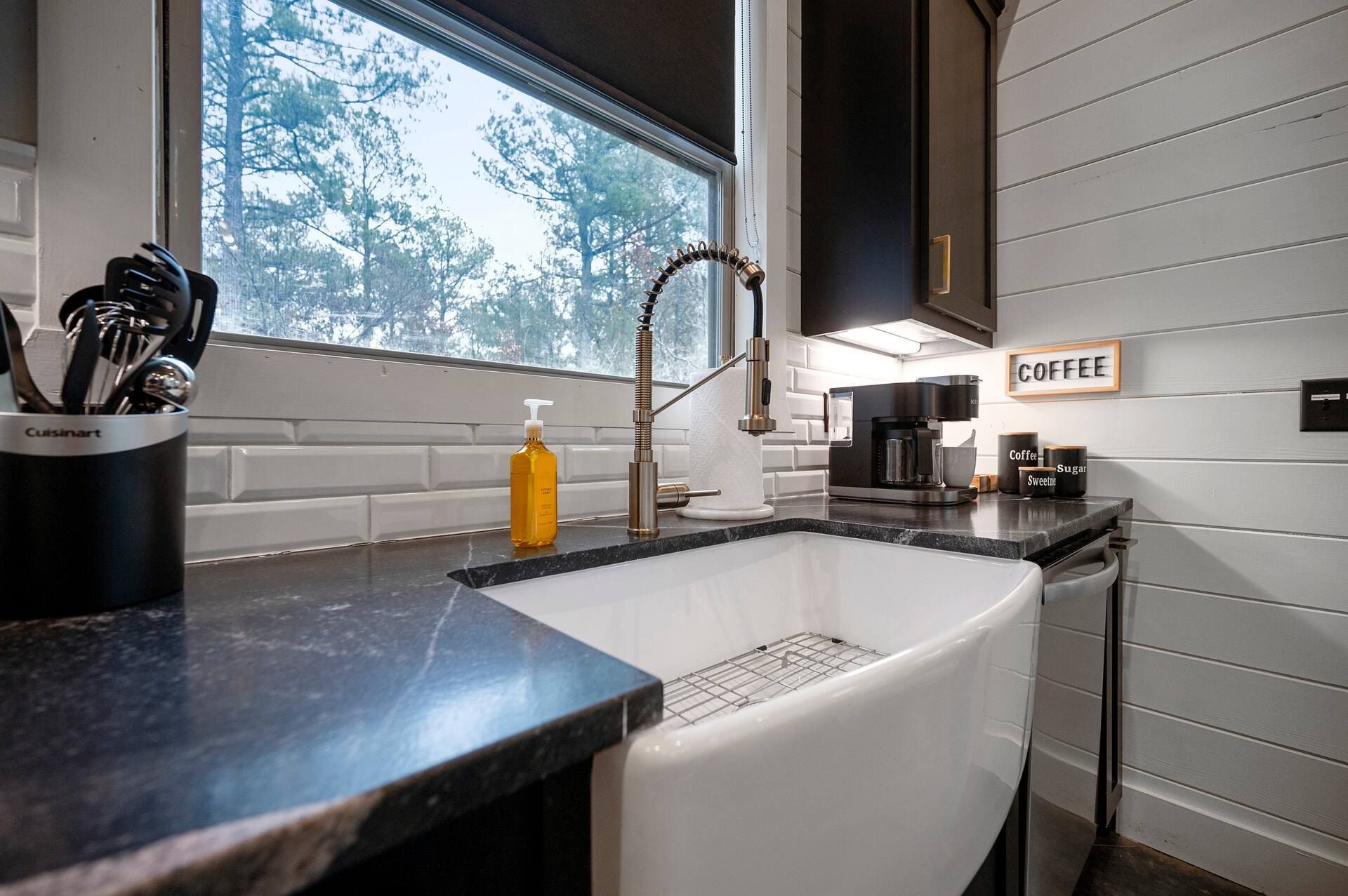 White farmhouse sink with a gold spring faucet under a window into the woods