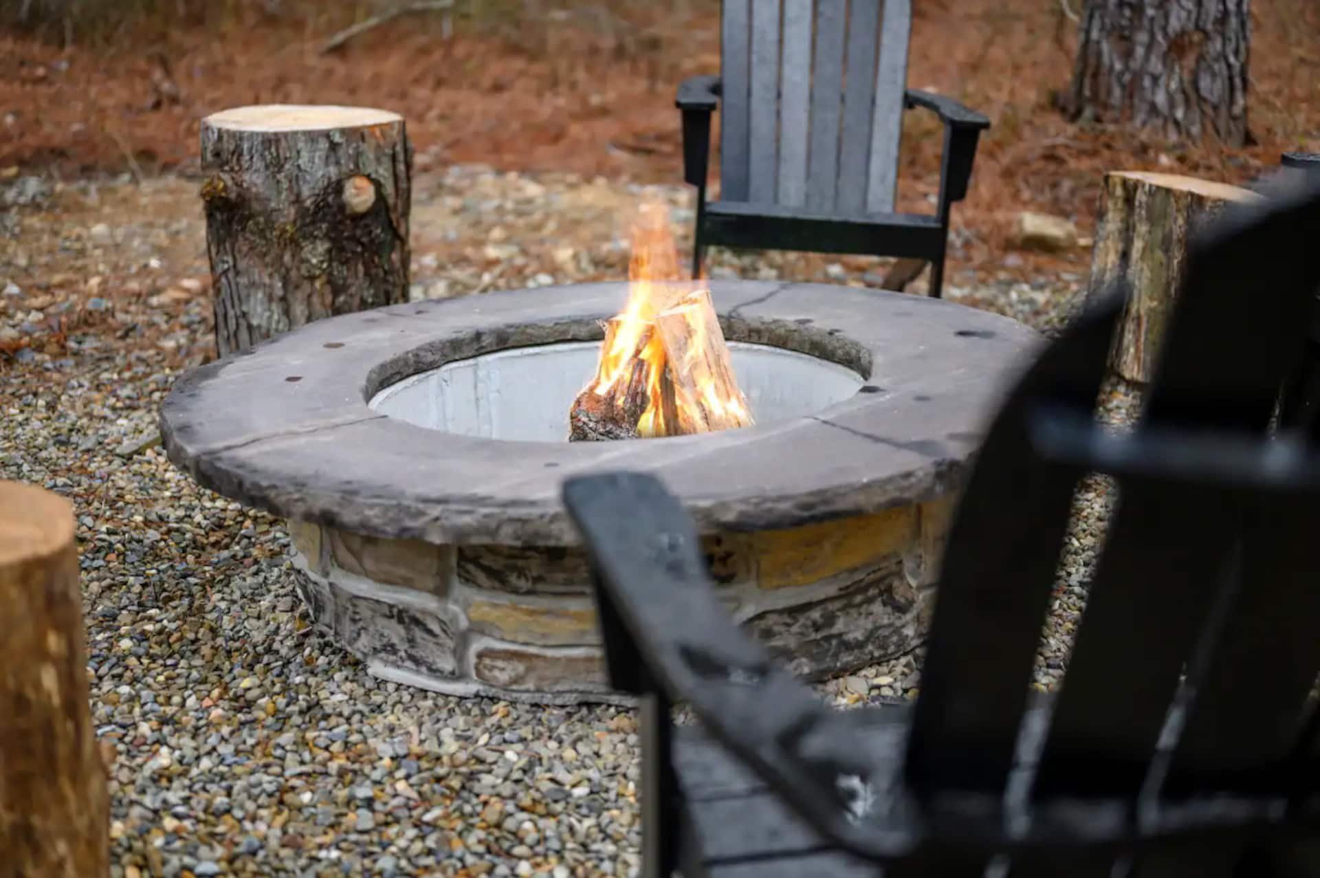 Stone fire pit burning with log stools and black Adirondack chairs on the gravel