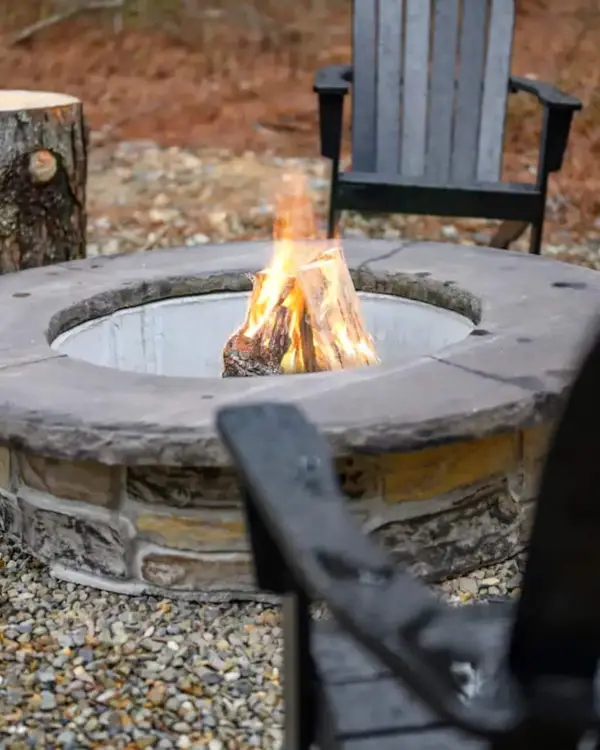 Stone fire pit burning with log stools and black Adirondack chairs on the gravel