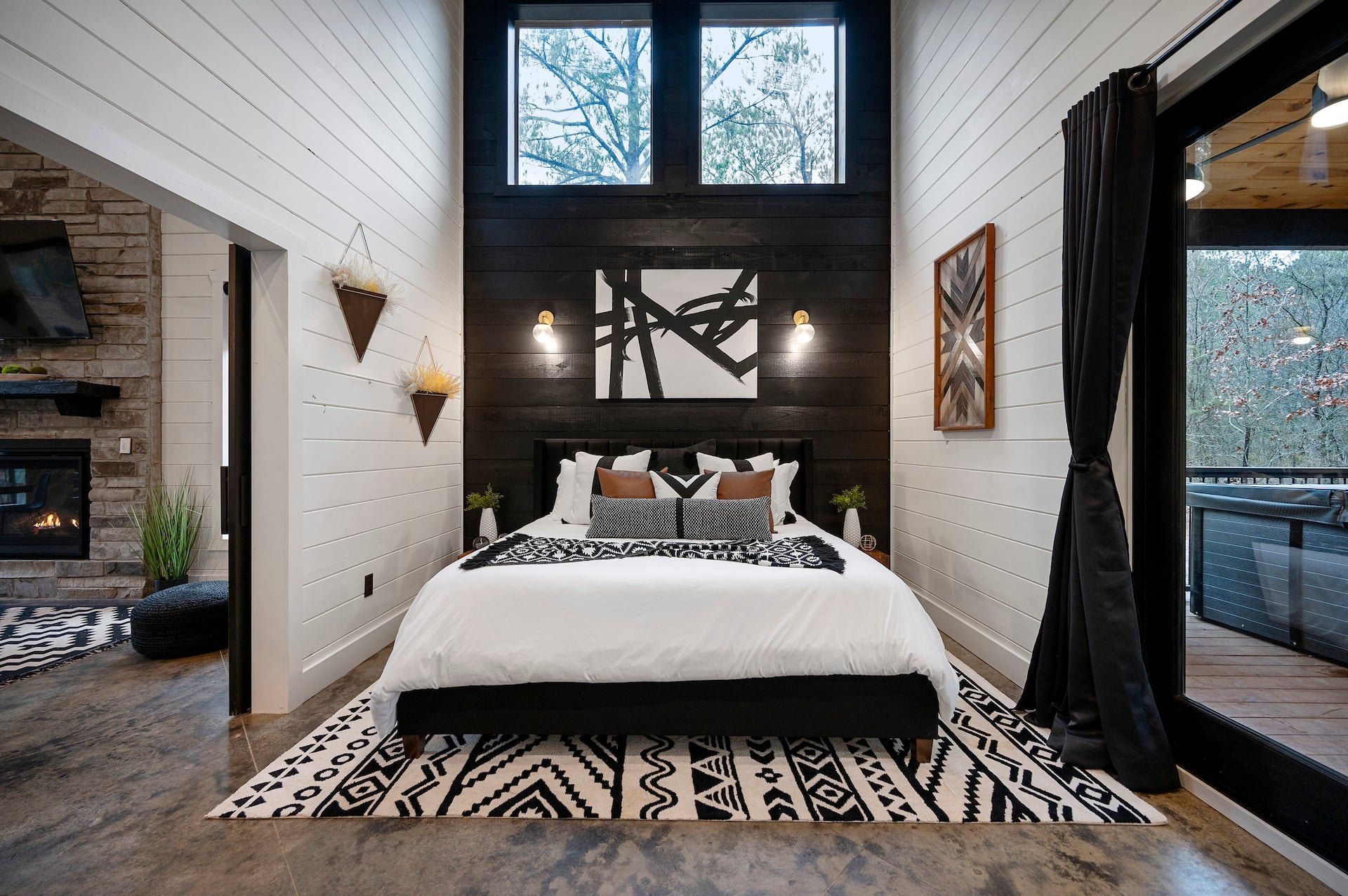 King bedroom seen from the doorway, black accent wall, geometric rug and tall windows to the pines