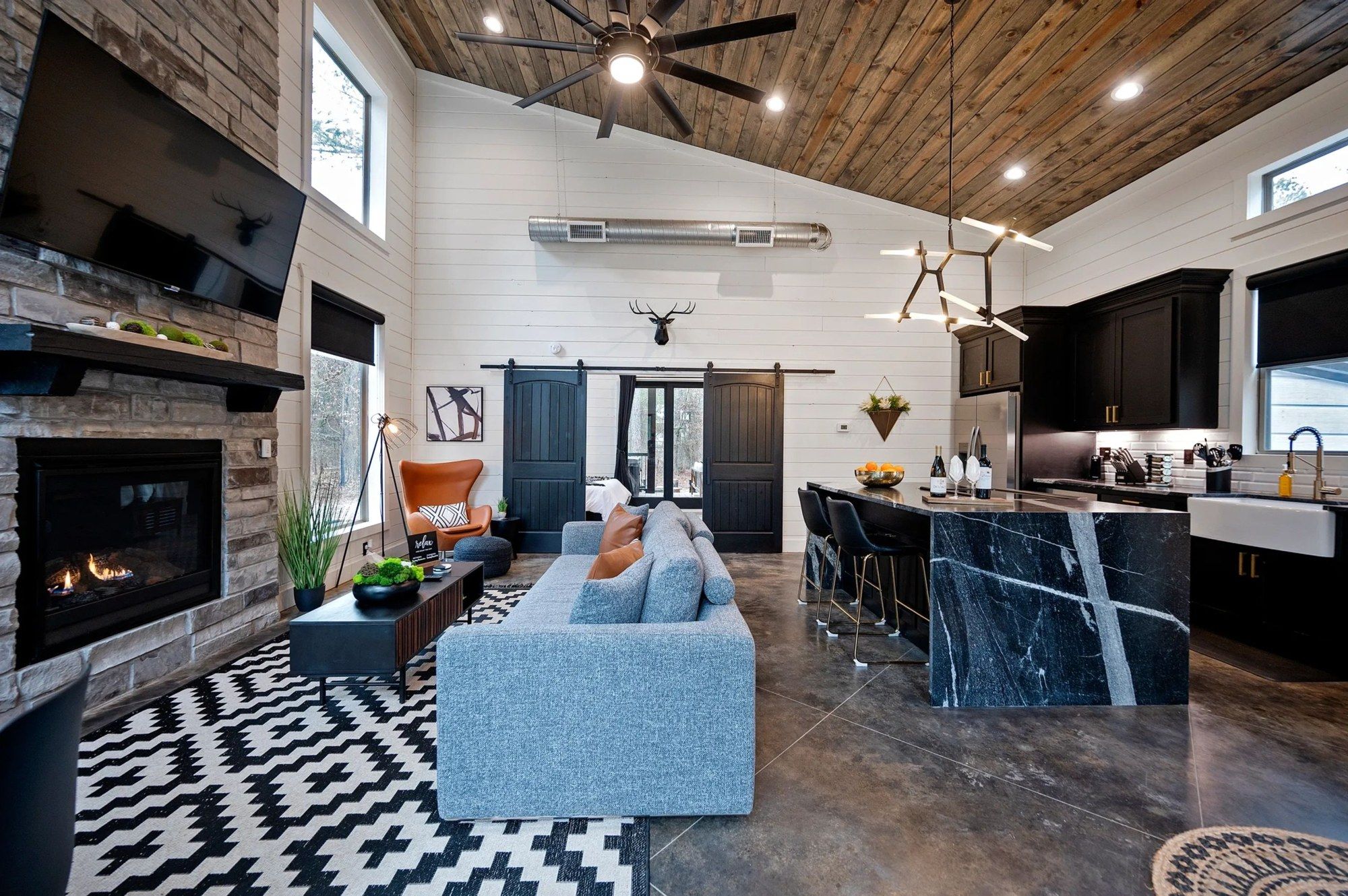 Living room with stone fireplace, blue sofa, barn door and the open kitchen under a vaulted wood ceiling