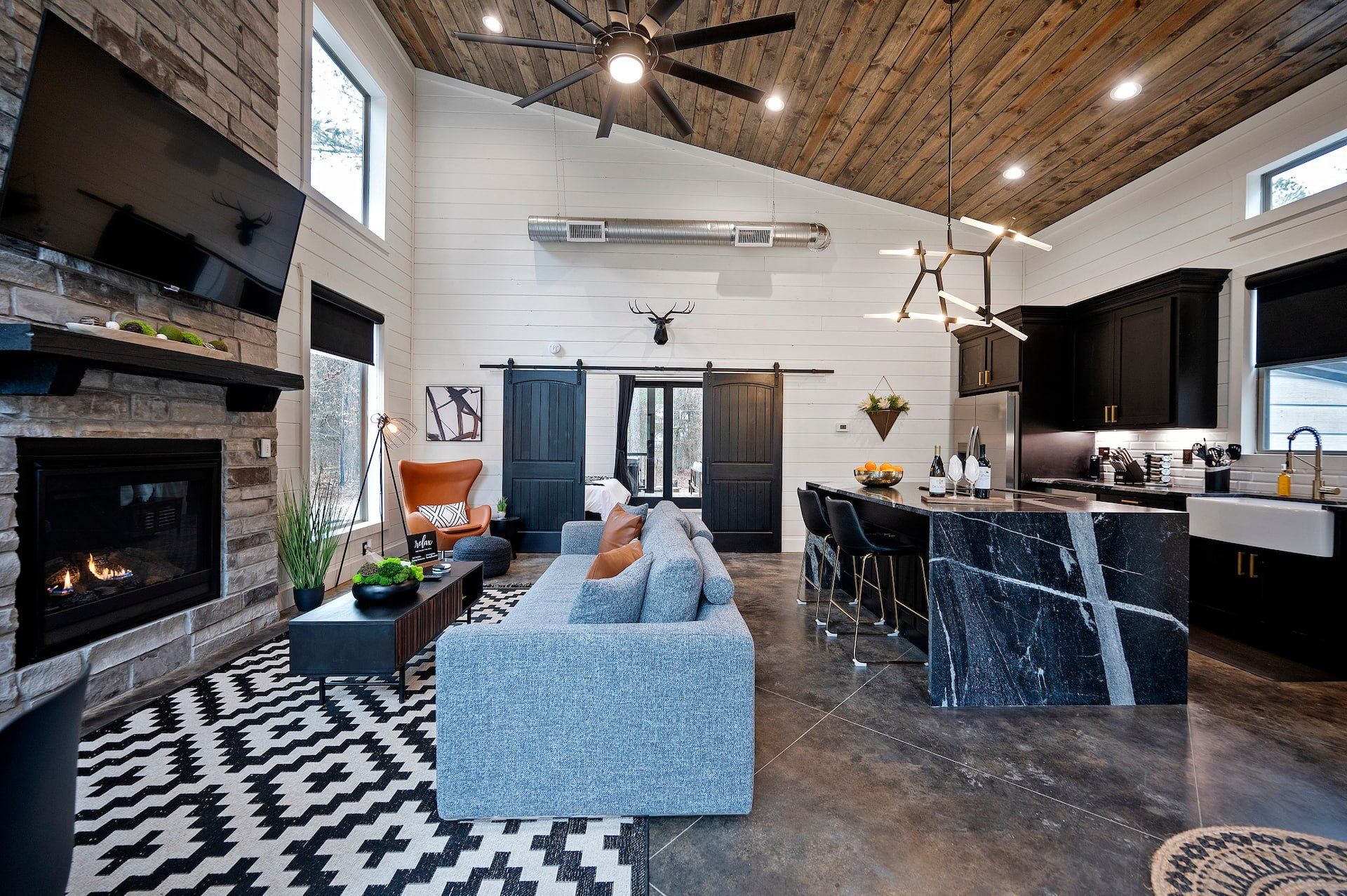 Open living room and kitchen under the vaulted wood ceiling, with the stone fireplace, blue sofa and black island