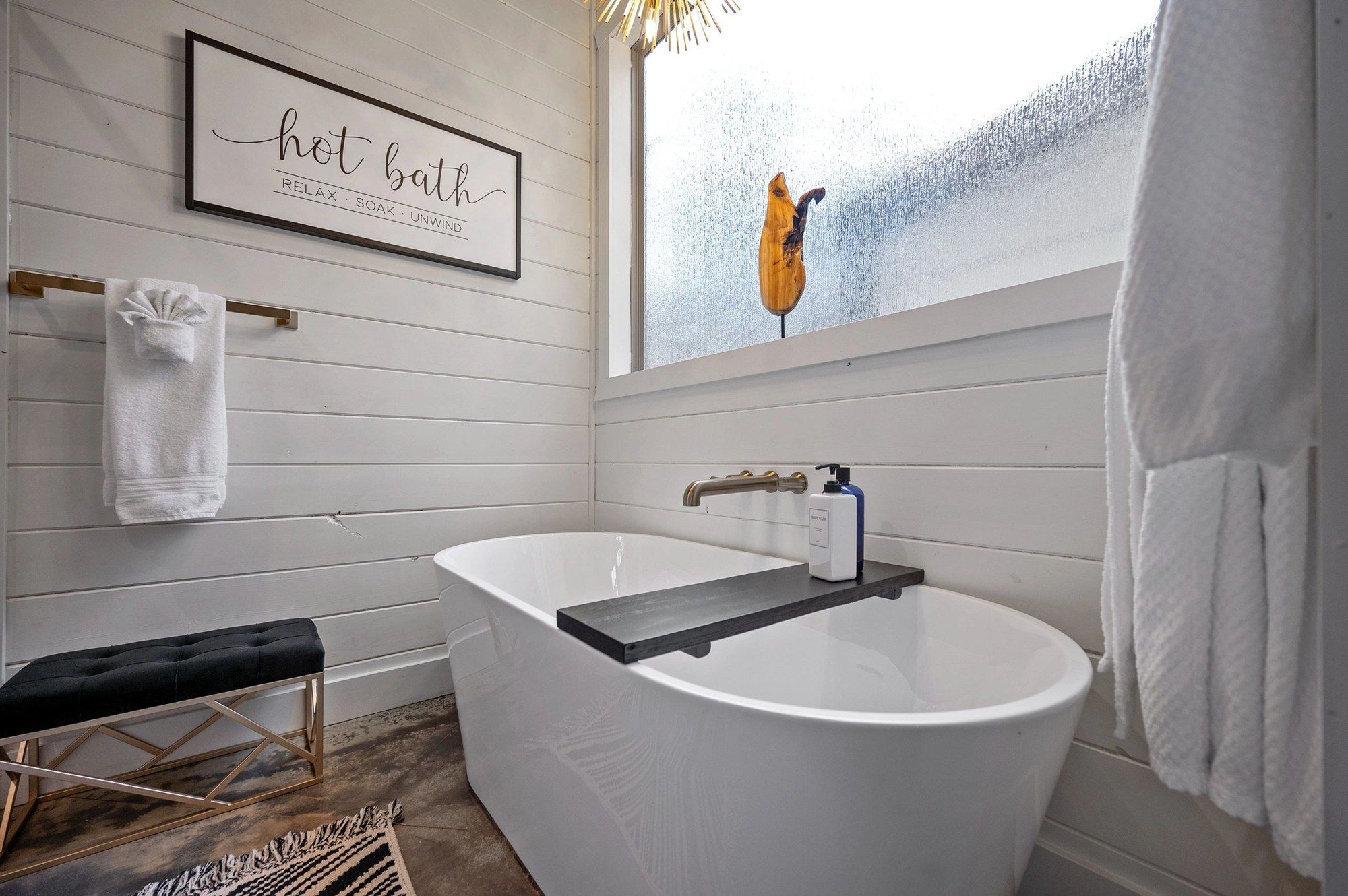 Freestanding soaking tub below a frosted window with a hot bath sign and a gold towel rail