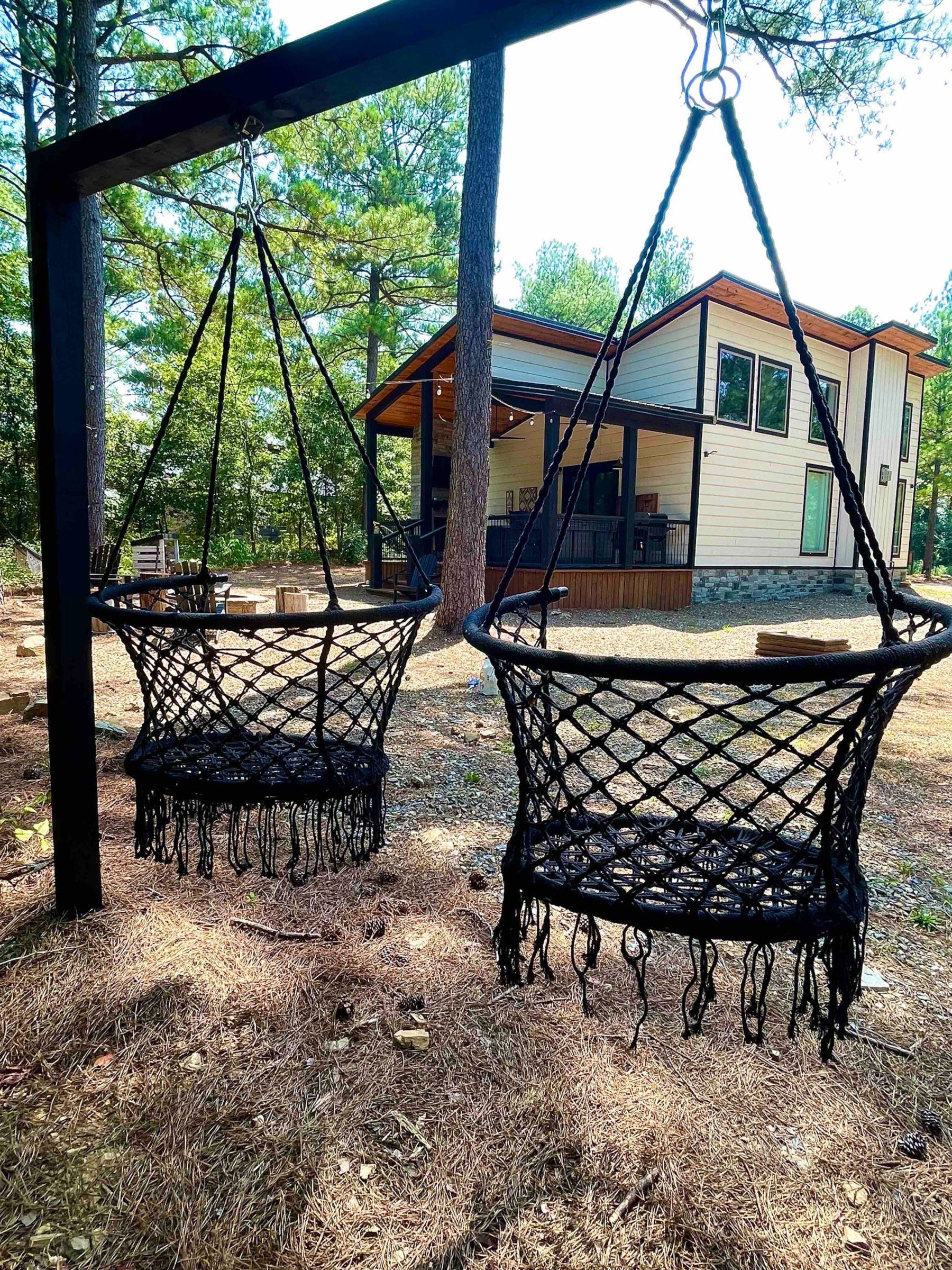 Two black macrame swing chairs on a frame under the pines with the cabin and deck behind